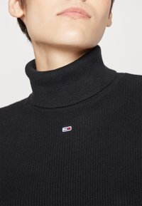 Maglione a collo alto nero con un design aderente. Presenta un piccolo logo ricamato sul petto in rosso, bianco e blu.