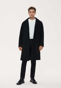 Long black coat made of wool with a notched collar, two buttons, and side pockets, paired with a white shirt and black pants.