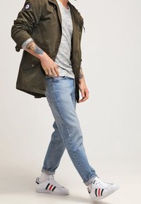 Olivgrön lättviktsjacka med en rund patch, grårandig t-shirt, ljusblå jeans och vita sneakers med röda och marinblå ränder.