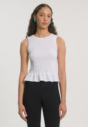 Young woman with curly hair wears sleeveless white textured top with ruffle hem, black pants, and blue hoop earrings against plain background.