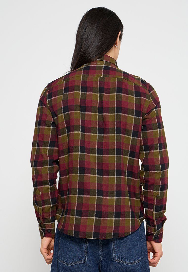 Personne avec de longs cheveux noirs portant une chemise à manches longues à carreaux rouge, olive et noir et un jean bleu, de dos contre un fond blanc.