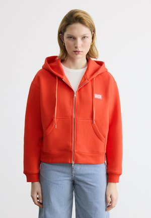 Jonge vrouw draagt een felrode hoodie met rits en voorzakken en gestreepte blauwe broek, staand tegen een effen witte achtergrond.