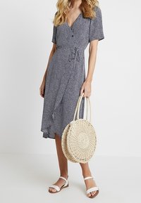 Blau-weiß gemustertes Wickelkleid mit kurzen Ärmeln, V-Ausschnitt und gebundenem Taillengürtel, kombiniert mit einer runden, geflochtenen Strohtasche und weißen Sandalen.