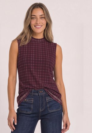Femme souriante portant un haut sans manches à carreaux bordeaux rentré dans un jean bleu foncé, sur un fond uni clair.