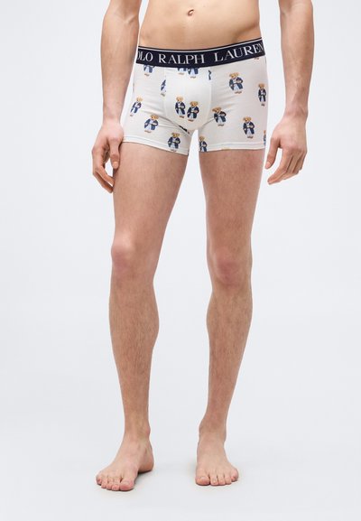 Άνδρας μοντ έλο φοράει λευκά εσώρουχα τύπου μποξεράκια με μπλε σκούρο λάστιχο μέσης με την επιγραφή "Polo Ralph Lauren" και σχέδιο αρκουδάκι, στέκεται ξυπόλητος σε λευκό φόντο.