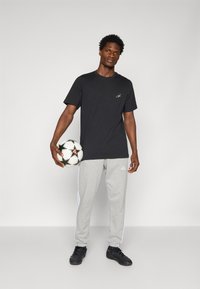 Sort kortærmet t-shirt med logo, grå sweatpants med hvide striber, holder en fodbold, sorte athleticsko.