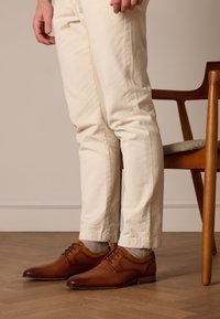 Bruine leren dresschoenen met een gestructureerde afwerking en vetersluiting, gecombineerd met lichtgekleurde broeken. Houten stoel op de achtergrond.