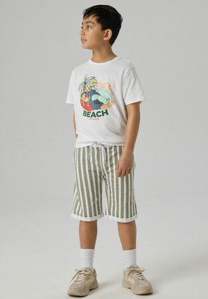 Junge steht, trägt ein weißes T-Shirt mit Strandgrafik und grün-weiß gestreifte Shorts, beige Turnschuhe und weiße Socken.