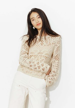 Jeune femme aux longs cheveux foncés portant un pull en crochet couleur crème et un pantalon blanc, debout devant un fond blanc.