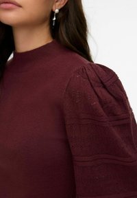 Pull bordeaux avec un col montant et des manches en dentelle bouffantes à texture stylisée. Agrémenté d'un corps en tricot lisse.