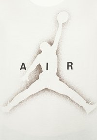 Vit t-shirt med en stor, brun grafik av en hoppande figur som håller en boll, med ordet "AIR" tryckt i svart under figuren.