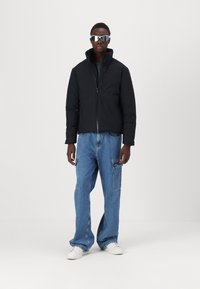 Black puffer jacket with a high collar, paired with light blue baggy jeans. The outfit is complemented by white sneakers and silver sunglasses.