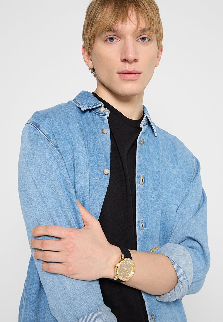 Camisa de mezclilla en azul claro con detalles de botones, llevada sobre una camiseta negra. El modelo lleva un reloj dorado con una correa negra en la muñeca.