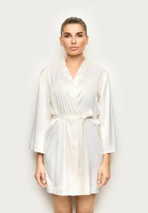 Femme debout de face portant un peignoir en satin blanc longueur genou, noué à la taille avec une ceinture, cheveux tirés en arrière, fond neutre.