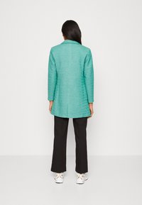 Türkisfarbener Blazer mit strukturiertem Finish, langen Ärmeln und Reverskragen, kombiniert mit schwarzen Hosen und weißen Sneakers.