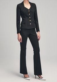 Woman wearing fitted black blazer with gold buttons, black flared pants, and white high heels against plain background.