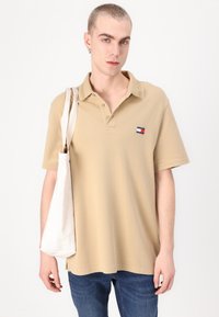 Beige polo skjorte med krage, tre-knaps lukning og lite logo. Modellen holder en lærremse tote bag. Har kortermet og avslappet passform.