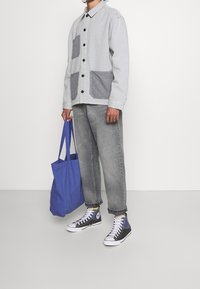 Szara kurtka zapinana na guziki z dwoma szarymi naszywanymi kieszeniami, w komplecie z szarymi dżinsami, białymi trampkami i niebieską torbą typu tote. Minimalistyczny design, luźny krój.