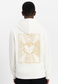 Baumwoll-Sweatshirt in Off-White mit einem großen goldenen Grafikdesign auf der Rückseite, das abstrakte Muster, ein Schmetterlingssymbol und strukturierte Linien zeigt.