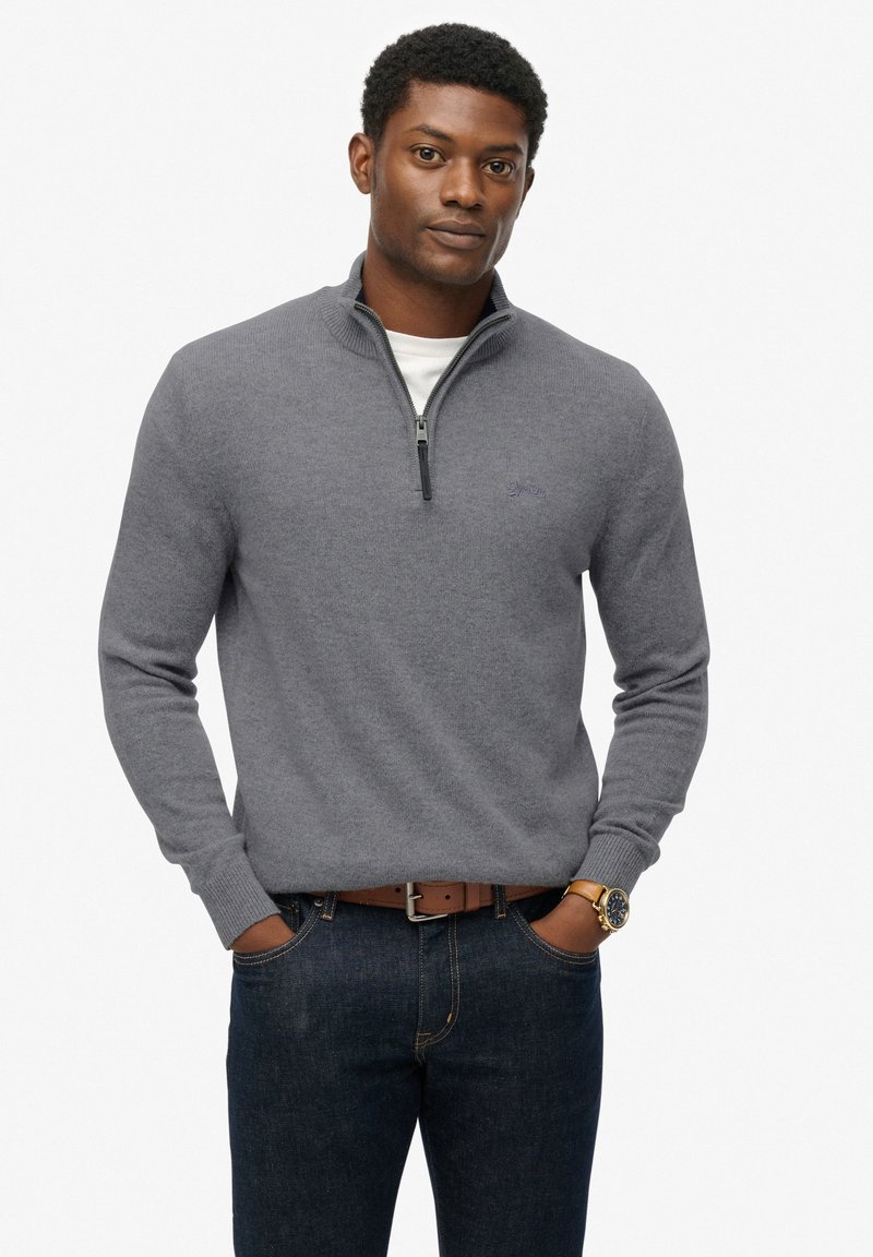 Pullover grigio a maglia con collo a zip e polsini a costine, indossato sopra una camicia bianca, abbinato a jeans scuri e una cintura marrone.
