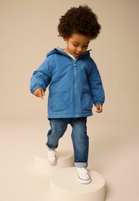 Lichtblauwe hooded jas met voorrits en twee zakken, gecombineerd met blauwe jeans en witte sneakers. Model staat op ronde platforms.