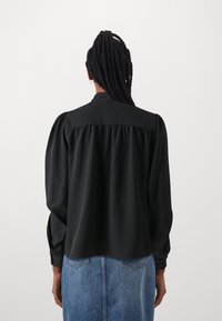 Svart bluse med lange ermer, puffede skuldrer, myk tekstur og knappekrage. Baksiden har rynker for en avslappet passform.