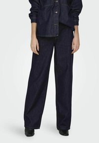 Pantalones de mezclilla oscuros de pierna ancha con cintura alta, que cuentan con un acabado limpio y detalles de costura mínimos, combinados con una chaqueta de camisa a juego.