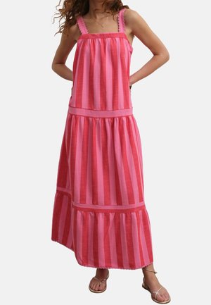 Vrouw draagt een mouwloze, roze en rode verticale gestreepte maxi-jurk met sandalen, staand met handen achter haar rug.