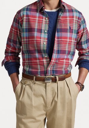 Homme portant une chemise à carreaux rouge et vert aux manches retroussées sur un pull bleu, pantalon beige avec ceinture marron et montre-bracelet. Mains dans les poches.