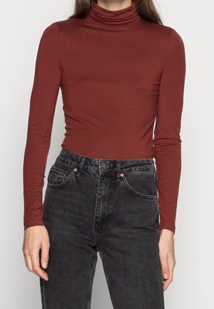 Kobieta ubrana w dopasowany, rdzawej barwy golf i czarne, wyprane jeansy z wysokim stanem, stojąca na jednolitym tle.