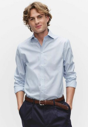 Nuori mies, jolla on vaaleanruskeat aaltoilevat hiukset, pukeutuneena vaaleansiniseen raidalliseen nappipaitaan, tummansinisiin housuihin ja ruskeaan punottuun vyöhön, kädet taskuissa.