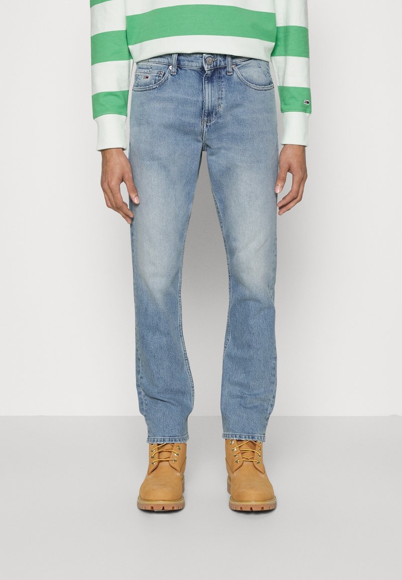 Vaqueros de mezclilla azul claro con corte recto y acentos desgastados, combinados con botas de cordones color marrón y una camisa de manga larga a rayas verdes.