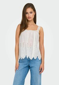 Canotta bianca in cotone con scollo quadrato, motivi di farfalle ricamati e orlo ondulato; abbinata a jeans azzurri chiari.