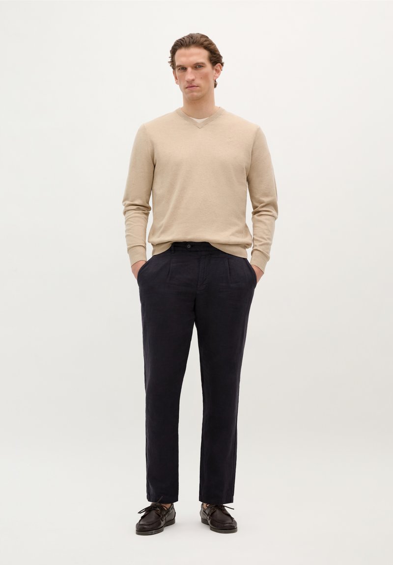 Beiger V-Ausschnitt-Pullover kombiniert mit dunklen marineblauen Leinenhosen und dunklen Schuhen. Das Outfit zeichnet sich durch ein entspanntes Design und eine glatte Textur aus.