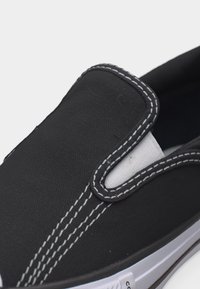 Czarny sportowy but wsuwany z białym przeszyciem, elastycznym bokiem i białą gumową podeszwą na jednolitym jasnym tle.