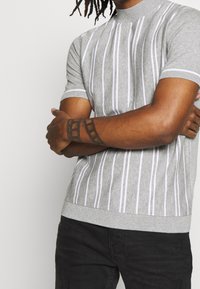 Pull gris en maille à manches courtes avec des rayures blanches verticales et un col montant. Comprend des poignets et un ourlet côtelés.