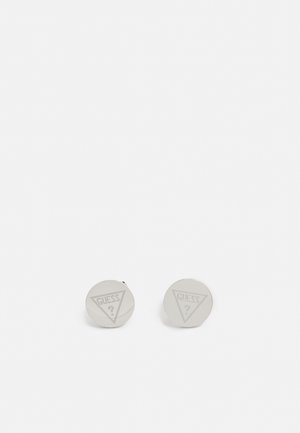 Okrągłe srebrne spinki do mankietów z wygrawerowanym logo GUESS w kształcie trójkąta oraz znakiem zapytania na jednolitym białym tle.