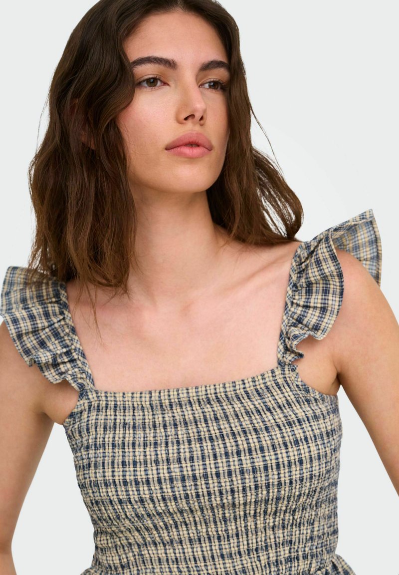 Jeune femme aux cheveux bruns ondulés portant une robe à carreaux bleue et beige sans manches avec des bretelles volantées, regardant légèrement sur le côté.