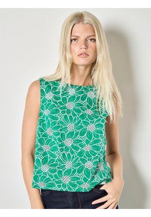 Mujer rubia con top verde sin mangas con un gran patrón floral blanco, parada con una mano en el bolsillo de mezclilla oscuro, fondo neutro.