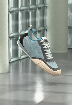 Hellblauer und grauer Sneaker mit weißen Schnürsenkeln und beiger Sohle, der drinnen auf einem polierten Betonboden in der Nähe einer Glasbausteinwand schwebt.