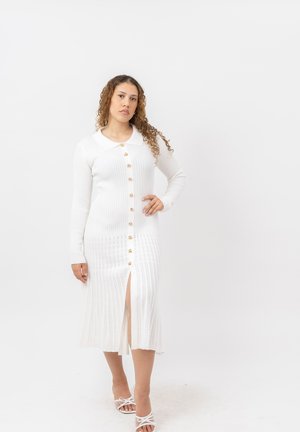 Frau mit lockigem Haar, die ein langes weißes, knöpfbares Kleid mit Kragen und weißen offenen Sandalen trägt, steht vor einem schlichten weißen Hintergrund.