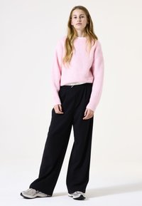 Roze geribbelde trui, zwarte wijde broek en grijze sneakers. Eenvoudige, losse pasvorm met een witte trekkoord in de taille. Plat, minimalistische achtergrond.