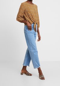 Braun gemusterte Bluse mit einem Knoten an der Taille, kombiniert mit hellblauen geraden Jeans und Sandalen mit Leopardenmuster.
