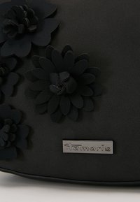 Schwarze Handtasche mit Stoff-Blumenverzierungen und einem metallischen Markenzeichen. Glatte Textur mit strukturiertem Design.