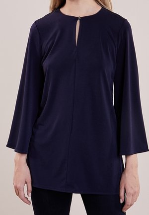 Kobieta w tunice z długim rękawem w ciemnoniebieskim kolorze, z małym wycięciem na przodzie i zapięciem na guzik.