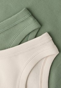 Deux pièces de sous-vêtements en coton côtelé, une vert sauge et une beige, superposées sur un tissu côtelé vert assorti en fond.