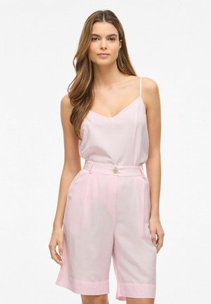 Kobieta z długimi brązowymi włosami, ubrana w jasnoróżowy top bez rękawów i różowe szorty z wysokim stanem, stojąca na tle jednolitego białego tła.