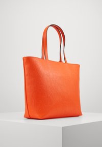 Pomarańczowa torba na ramię wykonana z teksturowanego materiału z wytłoczonym logo. Podwójne uchwyty w kontrastującym czarnym kolorze. Prostokątny kształt.