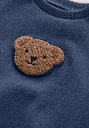 Navy blue sweatshirt featuring a textured, plush bear face patch in light brown with embroidered details. Ribbed neckline. Soft fabric texture.