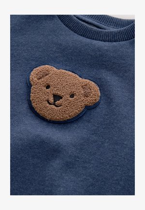 Tamsiai mėlynas džemperis su tekstūruotu, pūkuotu meškos veido pleistru šviesiai rudos spalvos su siuvinėtomis detalėmis. Ribinė apykaklė. Minkšta audinio tekstūra.